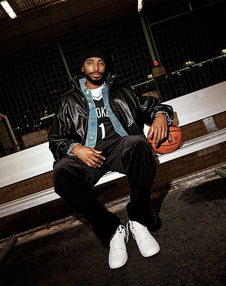Athletic man in a beanie and sports jersey sits on a bench with a basketball in an outdoor urban setting at night.