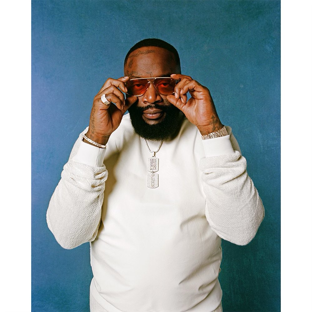 Portrait of Rick Ross wearing sunglasses and a white sweater in front of a mottled blue background, showcasing a celebrity lifestyle shoot.