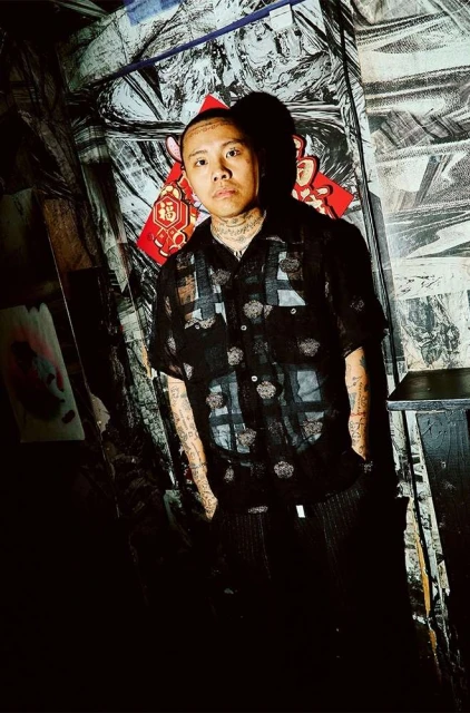 Portrait of a young man with tattoos, wearing a sheer button-down shirt, standing indoors against a textured, patterned background.