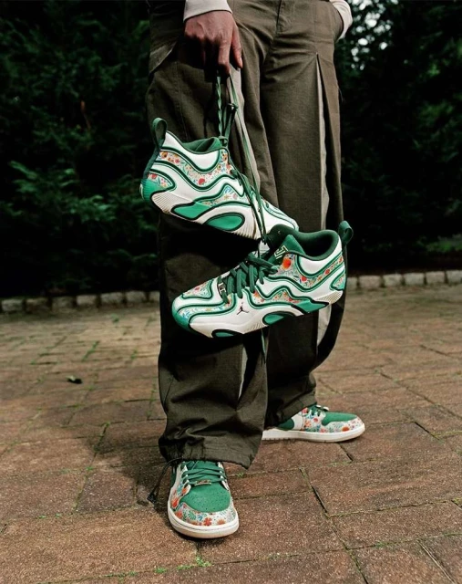 Person in olive pants displaying patterned green and white sneakers outdoors on a brick surface, with a sneaker pair hanging from the hand.