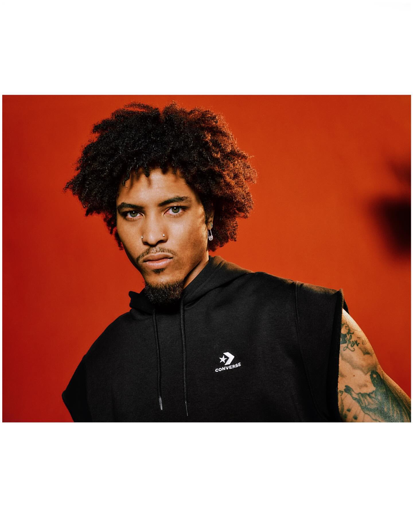 Portrait of a young adult man with curly hair, wearing a black sleeveless hoodie with a Converse logo against an orange background.