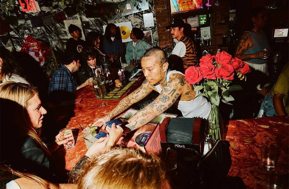 Patrons at a dimly lit bar, including an adult with many tattoos, red roses in a vase, bar counter, drinks, and other patrons in the background.