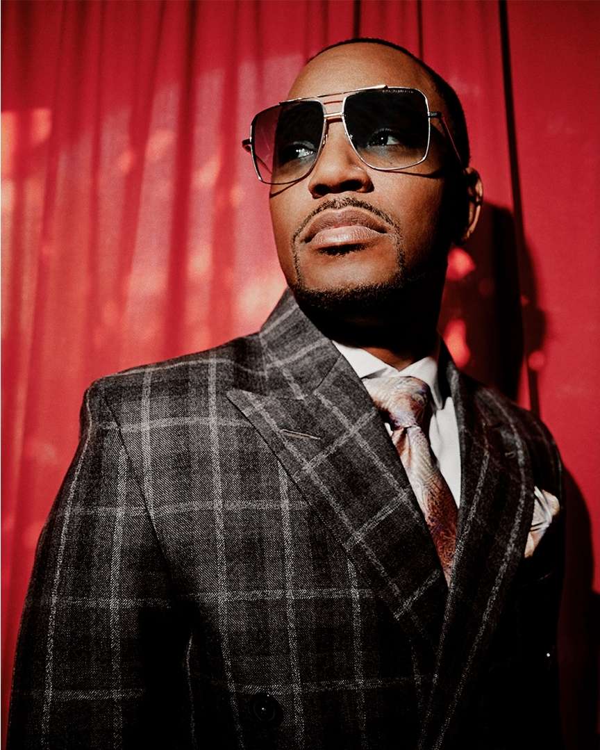 Portrait of an adult male wearing sunglasses and a grey plaid suit in front of a red curtain, looking off-camera.