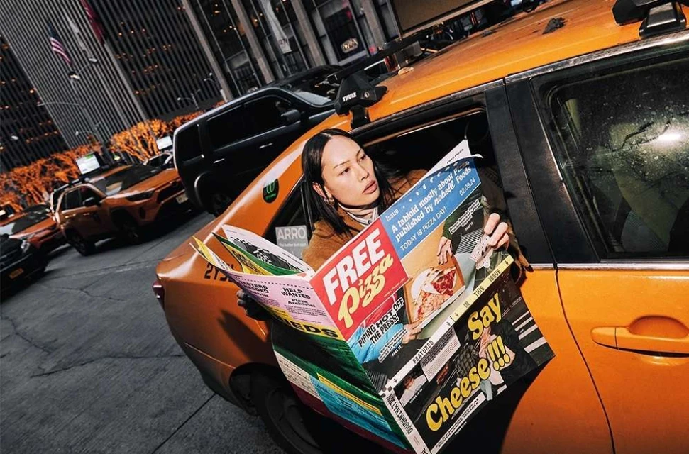 Woman reads 'Free Pizza' magazine in a taxi against a backdrop of city buildings and traffic, creating an urban editorial scene.