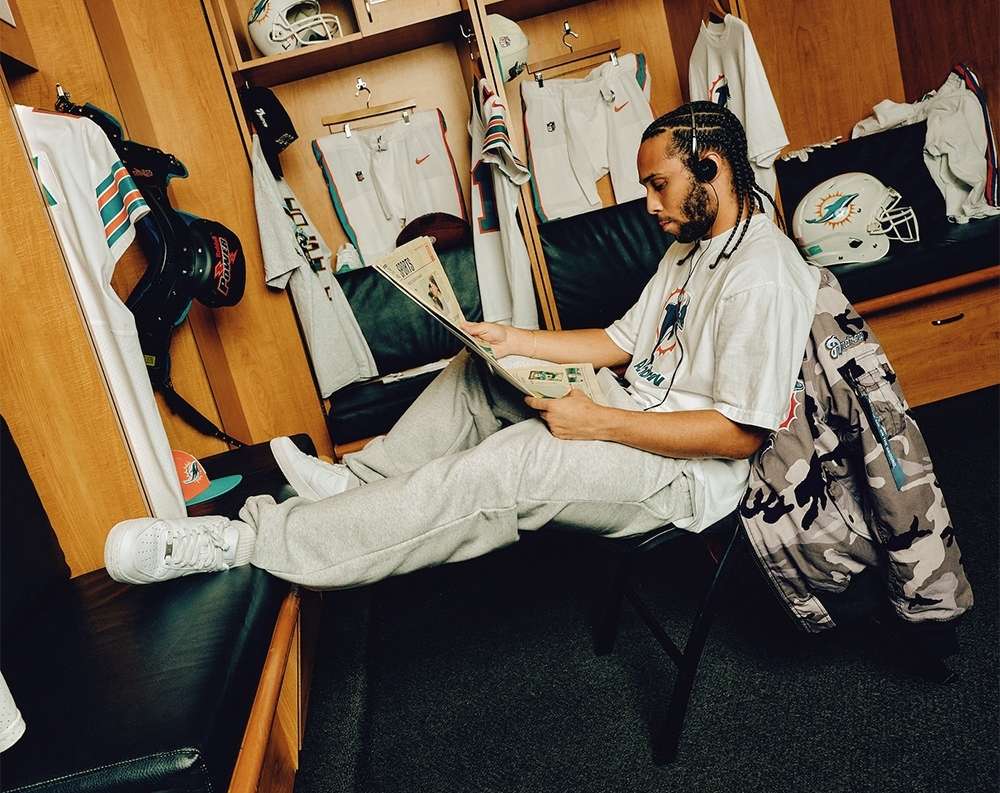 Man in locker room wearing Miami Dolphins apparel, sitting and reading. Lockers contain football gear and clothing with team logos.