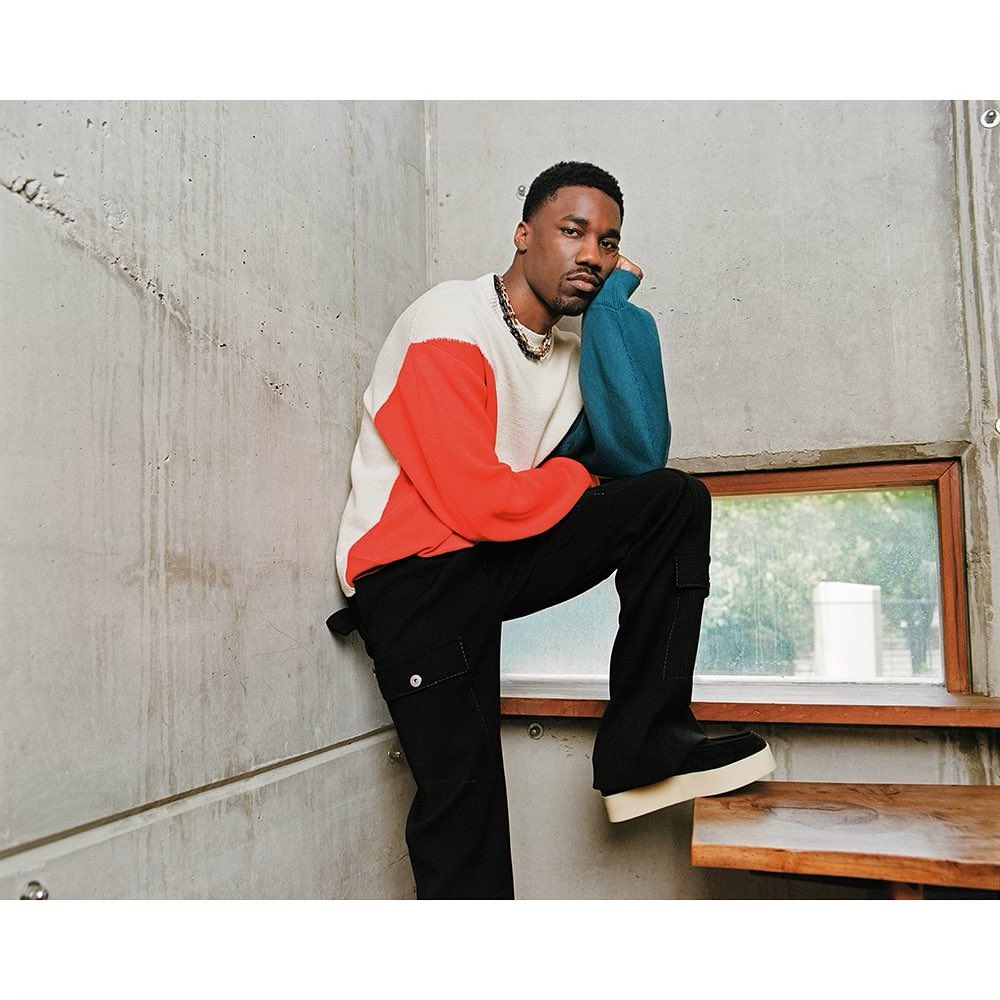 Stylish man in a color-blocked sweater, black pants, and platform shoes poses against a concrete wall and wooden stairs indoors.