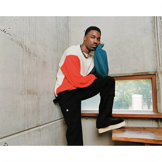 Stylish man in a color-blocked sweater, black pants, and platform shoes poses against a concrete wall and wooden stairs indoors.