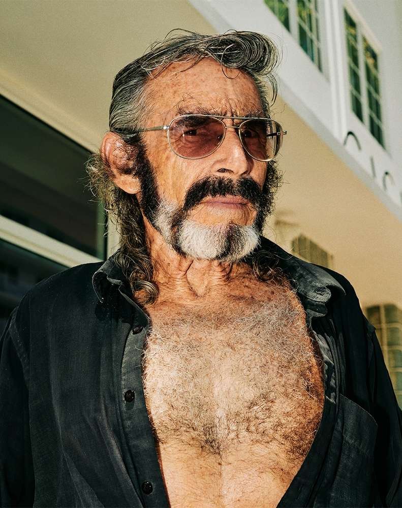 Close-up portrait of an older adult man with a beard, sunglasses, and an open buttoned shirt against an outdoor building backdrop.