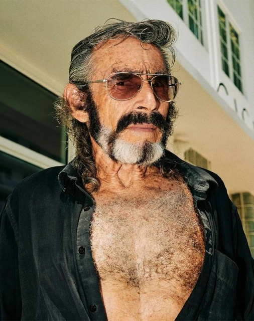 Close-up portrait of an older adult man with a beard, sunglasses, and an open buttoned shirt against an outdoor building backdrop.