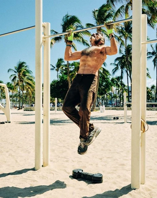 Man performs pull-up on outdoor bar at the beach with palm trees and blue sky in the background. Fitness concept in sunlight.