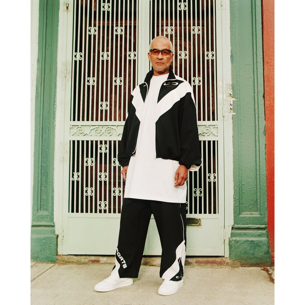 Full-shot portrait of an adult male wearing a black and white tracksuit in front of a green and white door in daylight.