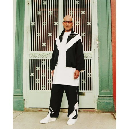 Full-shot portrait of an adult male wearing a black and white tracksuit in front of a green and white door in daylight.