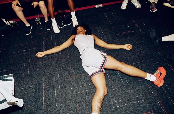 Young athlete lying flat on a dark patterned carpeted floor surrounded by teammates standing nearby, photographed by David Cabrera