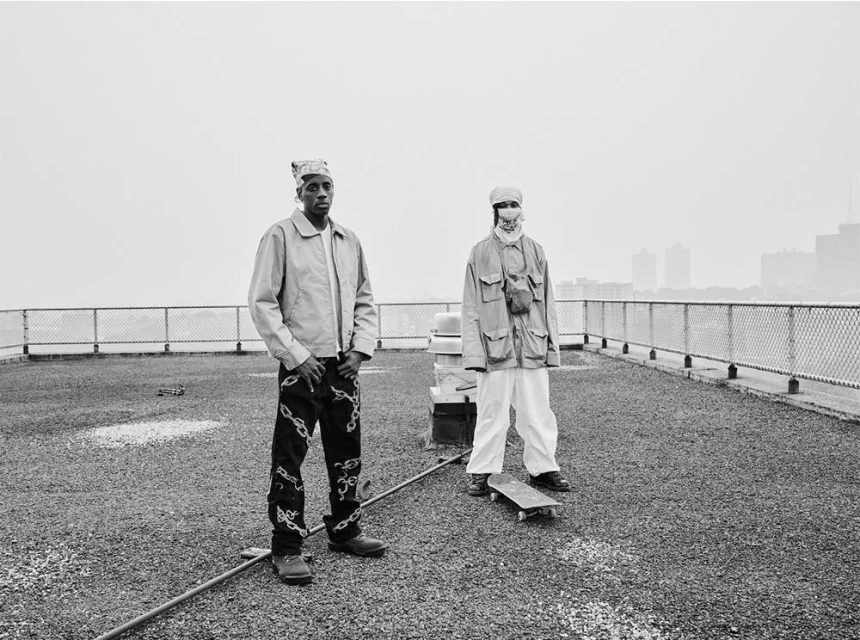 Two adults in jackets and bandanas stand on a rooftop with a skateboard, with city buildings in the distance against a light grey sky.