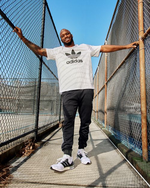 Athletic adult male in Adidas apparel stands in a walkway with arms outstretched, showcasing fitness and outdoor lifestyle fashion against a blue sky.