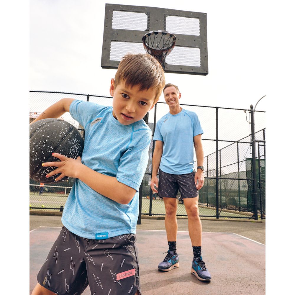A boy and adult male stand on a basketball court. The boy holds a basketball, and a backboard is visible in the background.