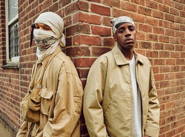 Two men standing against a red brick wall, wearing tan jackets and head coverings, photographed by David Cabrera.