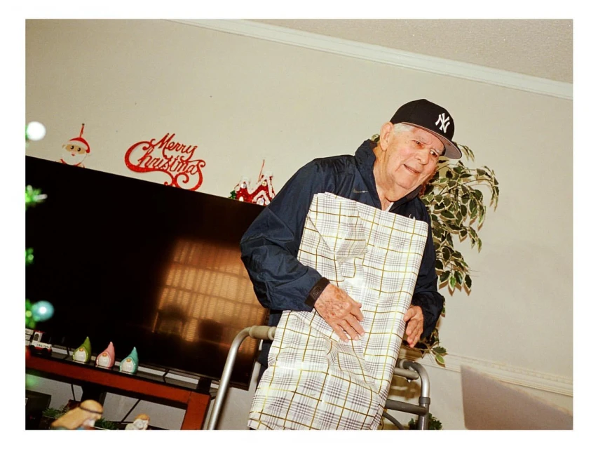 Elderly man in baseball cap holding a wrapped gift with a walker indoors, with Christmas decorations and a TV in the background.