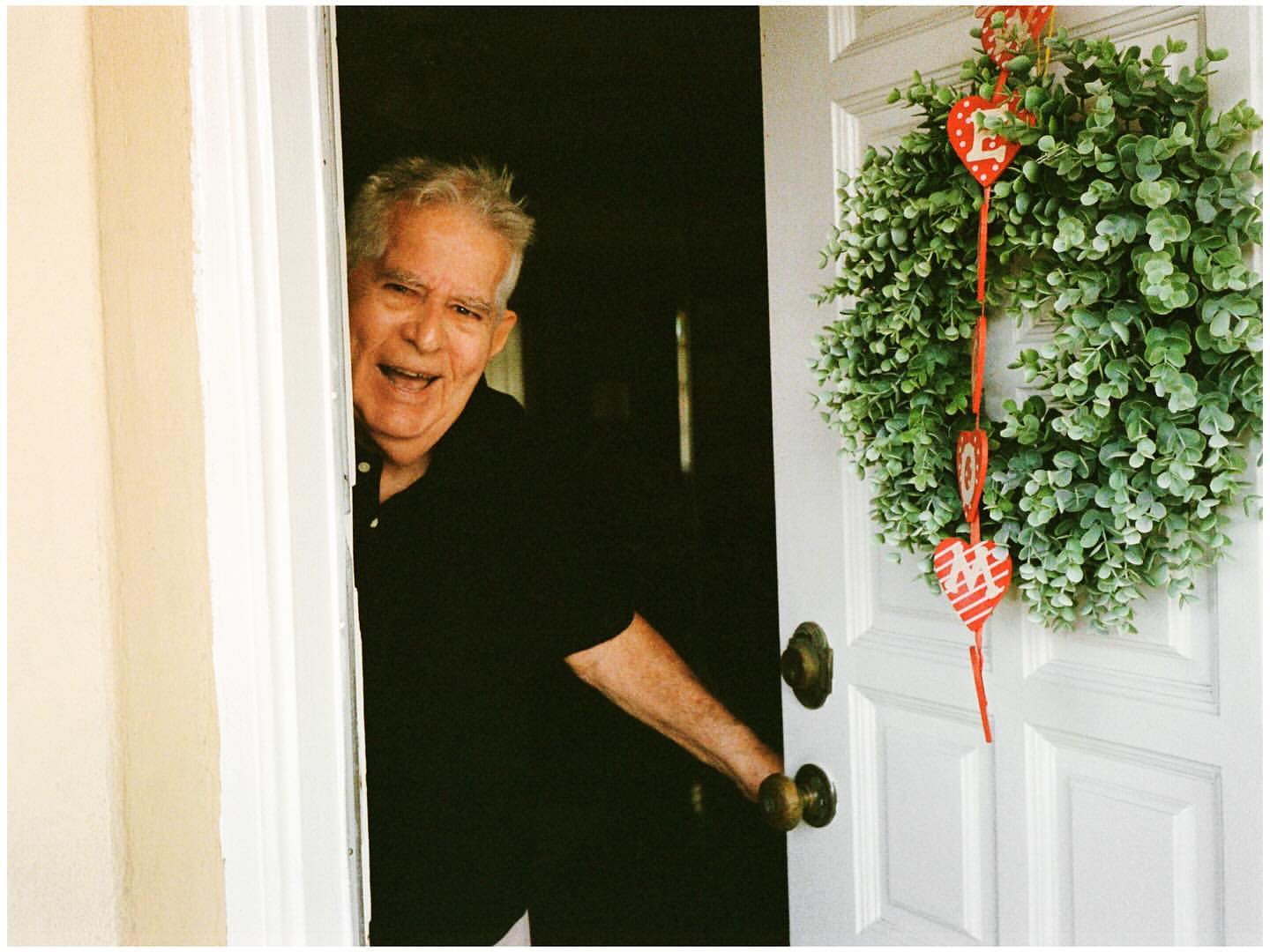 Smiling elderly man at doorway with holiday wreath. Dark shirt and white doorframe, with green and red heart-shaped wreath.