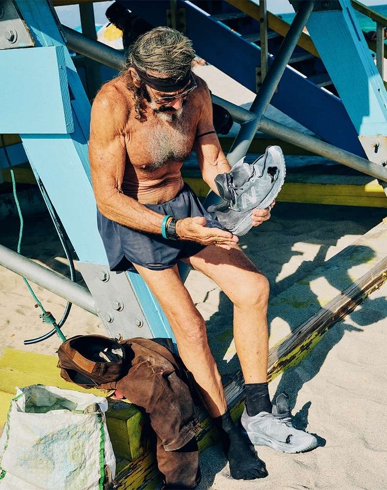 Older man with long gray hair sits outdoors, holding sneakers on beach sand in front of blue and white lifeguard tower.