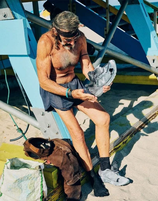 Older man with long gray hair sits outdoors, holding sneakers on beach sand in front of blue and white lifeguard tower.