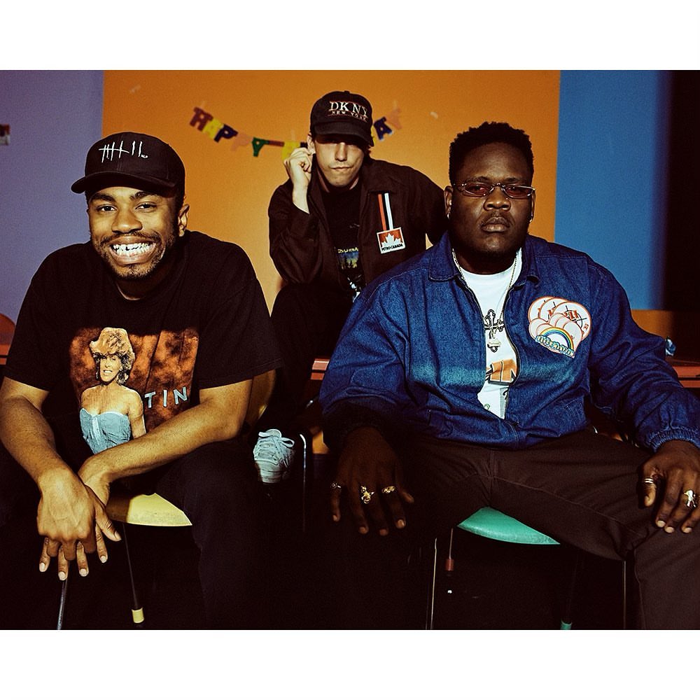 Three men pose indoors with colorful background. One wears a DKNY hat and a jacket with the Marlboro logo visible.