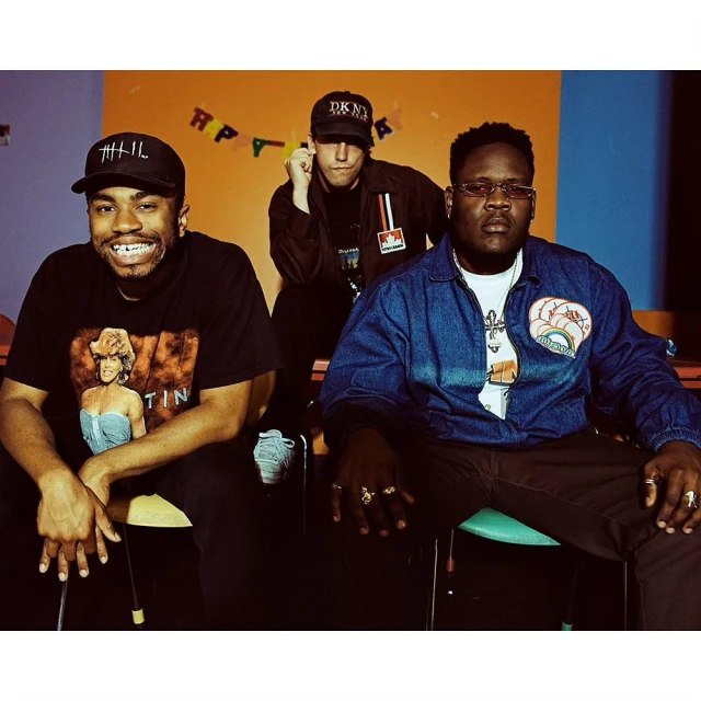 Three men pose indoors with colorful background. One wears a DKNY hat and a jacket with the Marlboro logo visible.