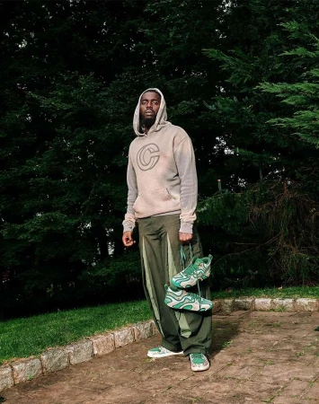 Black man wearing a grey hoodie and olive green pants holding green sneakers outdoors