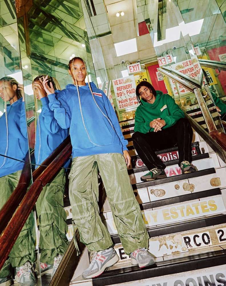 Two fashion models pose on a colorful staircase in hoodies and cargo pants, reflected in mirrored walls with visible signage.