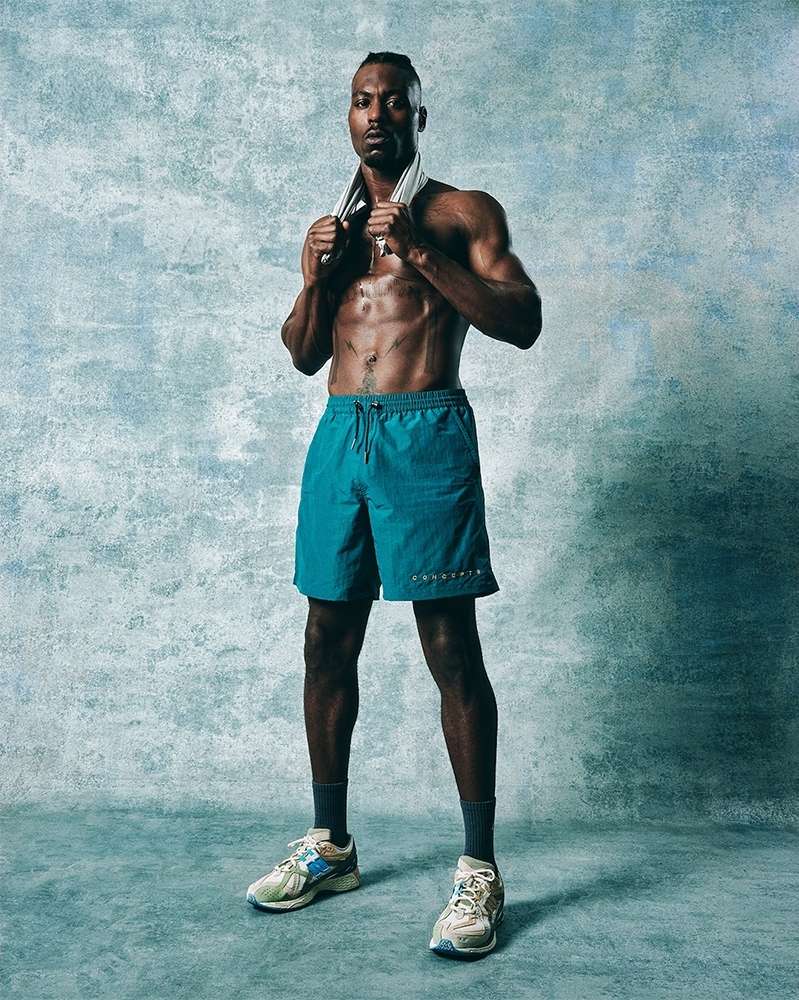 Athletic adult male stands with a towel around his neck in a full shot against a textured blue studio backdrop, wearing teal shorts and sneakers.