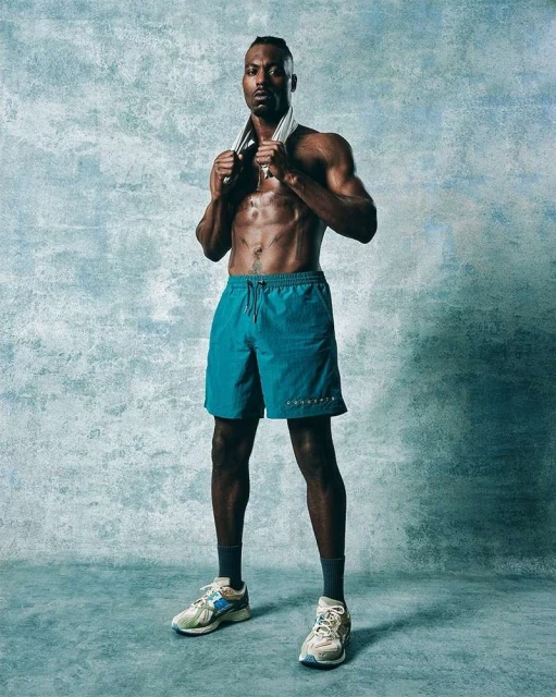Athletic adult male stands with a towel around his neck in a full shot against a textured blue studio backdrop, wearing teal shorts and sneakers.