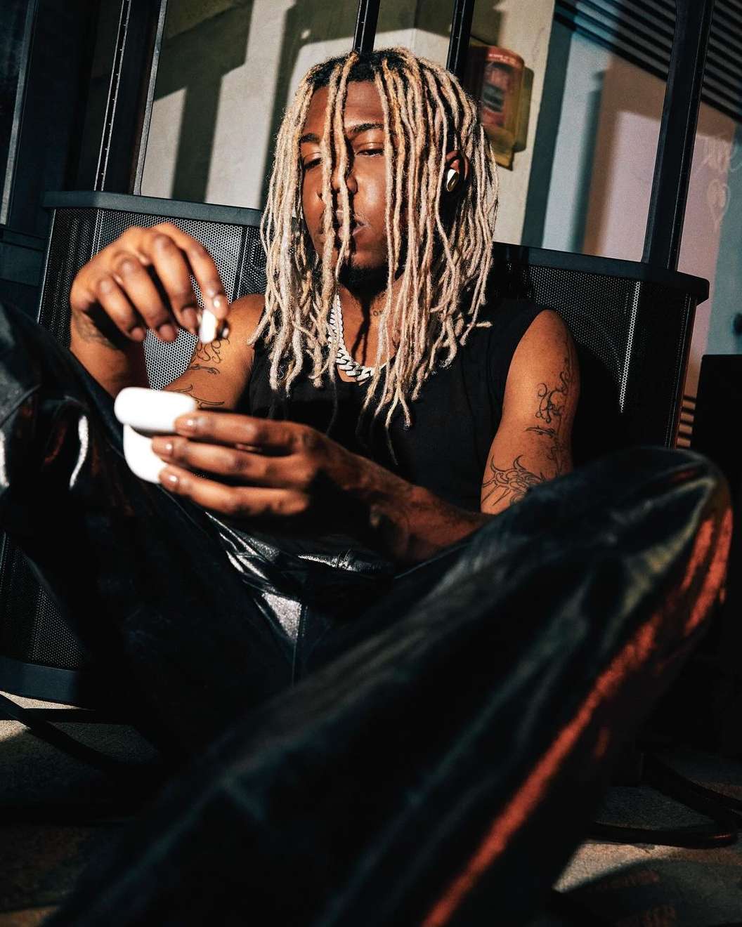 Young adult with dreadlocks sits indoors in black tank top and pants, holding earbuds case in hand, tattoos visible on arms.