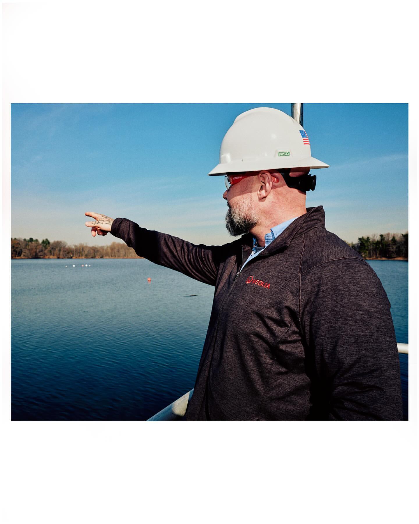 Adult male engineer wearing a hard hat points toward a lake surrounded by trees under a clear blue sky during the day.