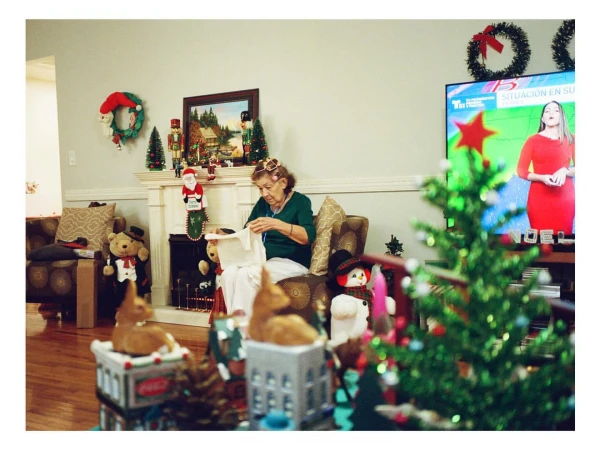 Elderly woman in green shirt opening a gift in a festive, decorated living room with a TV displaying a news broadcast.