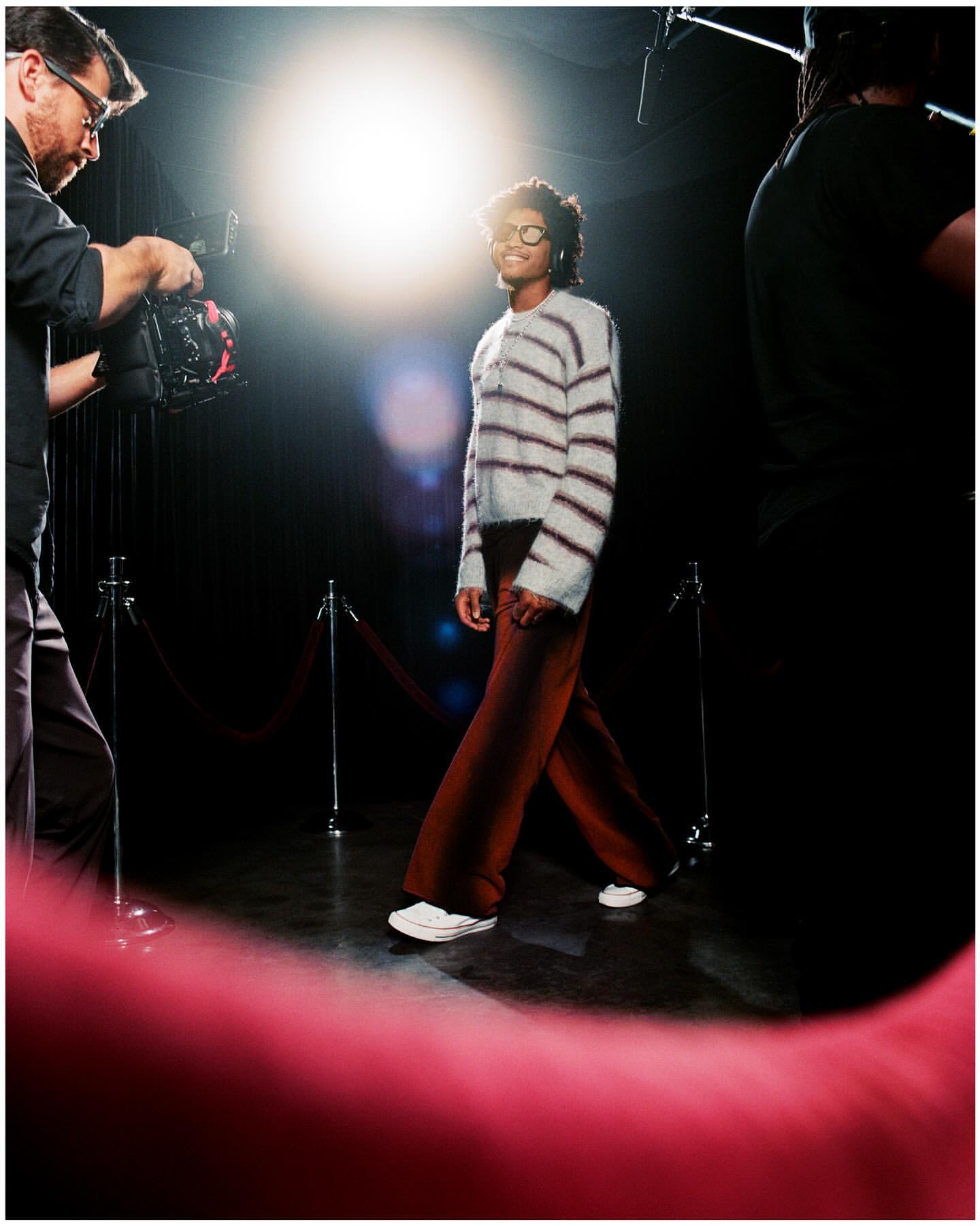 Woman walking on a red carpet in a striped sweater and brown pants, being filmed by a cameraman with studio lighting.