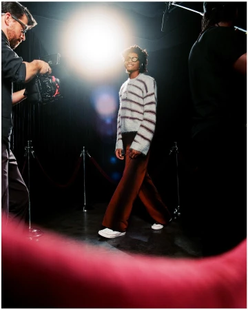 Woman walking on a red carpet in a striped sweater and brown pants, being filmed by a cameraman with studio lighting.