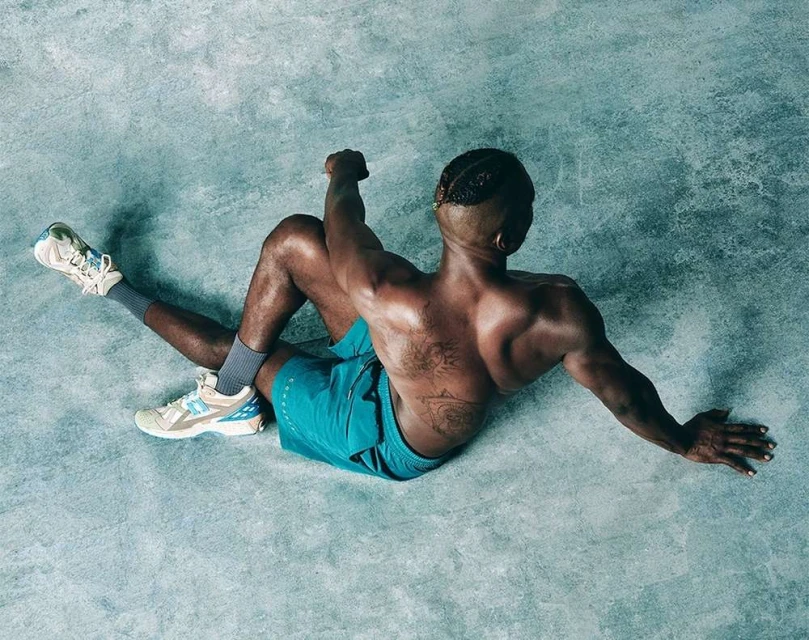 Overhead shot of a fit Black man in turquoise shorts and sneakers sitting on the ground in a spinal twist pose in a fitness studio.