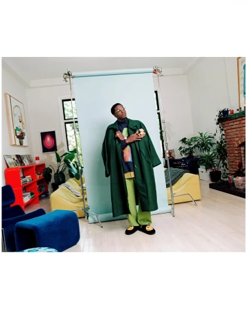 Man wearing a green coat and smiley face slippers standing in front of a light blue photo backdrop in a brightly lit interior space.