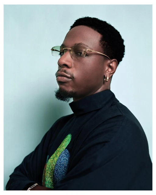 Portrait of a stylish adult Black man with dark hair, gold-rimmed glasses, and a black turtleneck sweater against a light blue background.