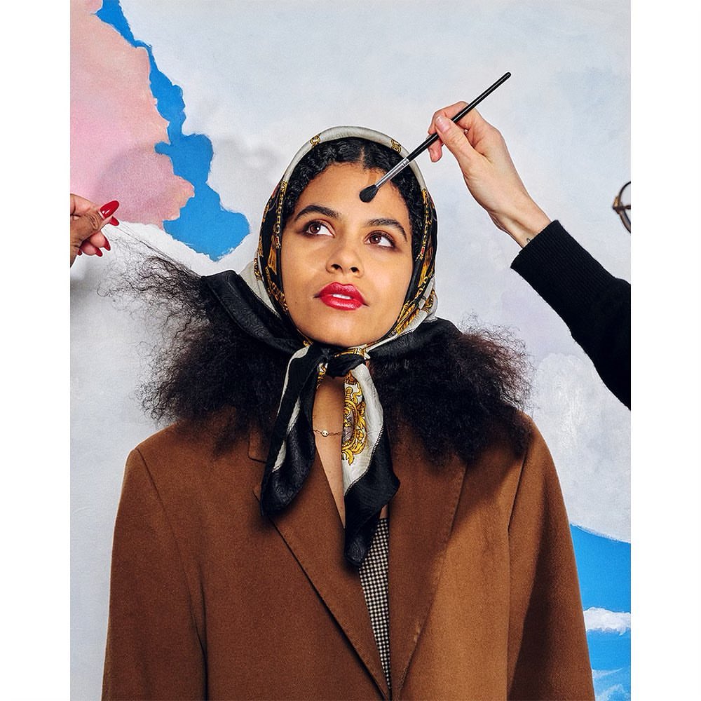 Brunette woman gets makeup applied by makeup artist in a beauty studio with an abstract painted backdrop; headscarf and brown jacket.