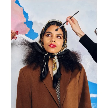 Brunette woman gets makeup applied by makeup artist in a beauty studio with an abstract painted backdrop; headscarf and brown jacket.