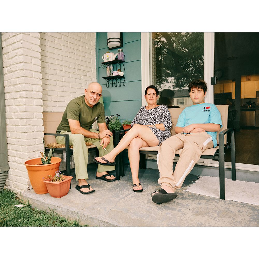 Family of three—adult male, adult female, and teen—sit on a porch with plants in pots; natural lighting.