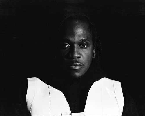 Monochrome portrait of an African American man with braids wearing a white vest and dark hoodie against a dark background.