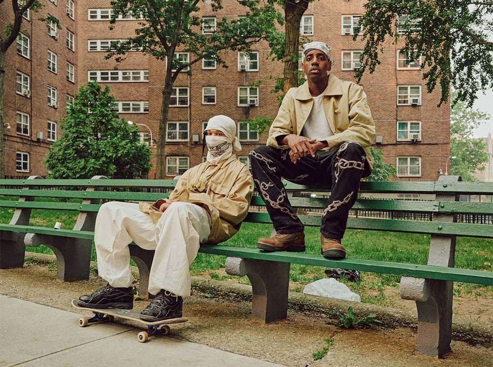 Two adult males sit on a park bench in front of an apartment building. One man sits on a skateboard. Urban setting, daytime.