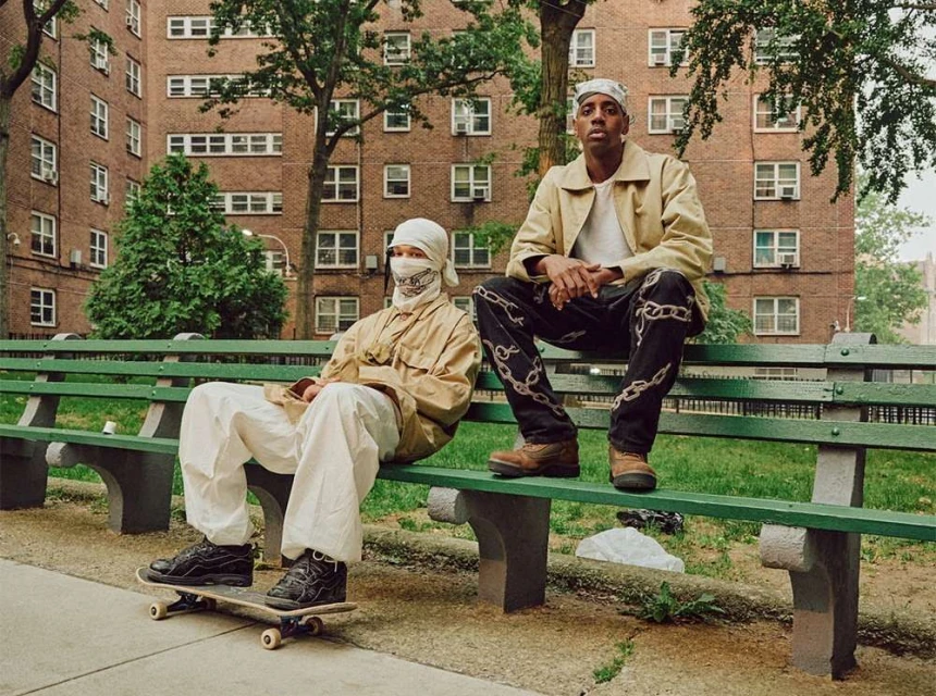 Two adult males sit on a park bench in front of an apartment building. One man sits on a skateboard. Urban setting, daytime.