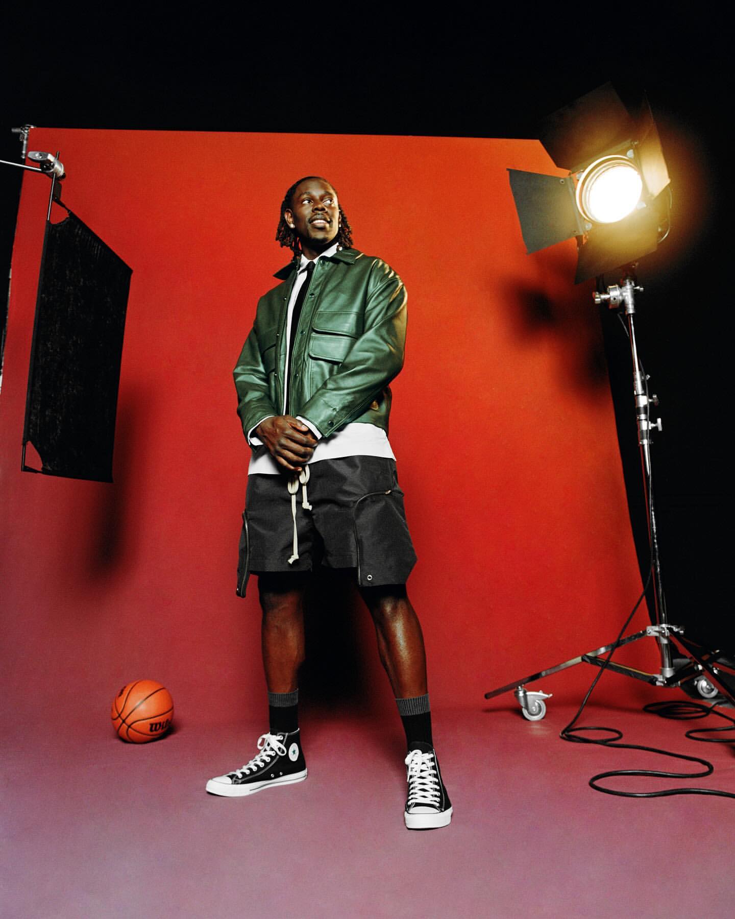Full studio shot of athletic adult male posing in green leather jacket, black shorts, and Converse sneakers. Basketball in foreground.