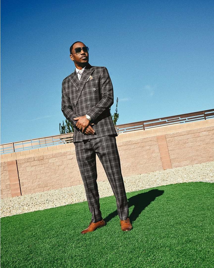An adult man in a plaid suit and sunglasses stands on artificial turf against a pale blue sky and a low, light brick wall.