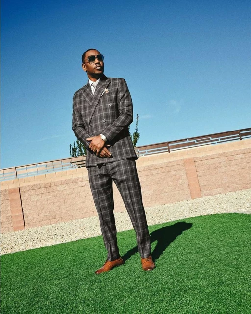 An adult man in a plaid suit and sunglasses stands on artificial turf against a pale blue sky and a low, light brick wall.