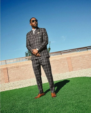 An adult man in a plaid suit and sunglasses stands on artificial turf against a pale blue sky and a low, light brick wall.