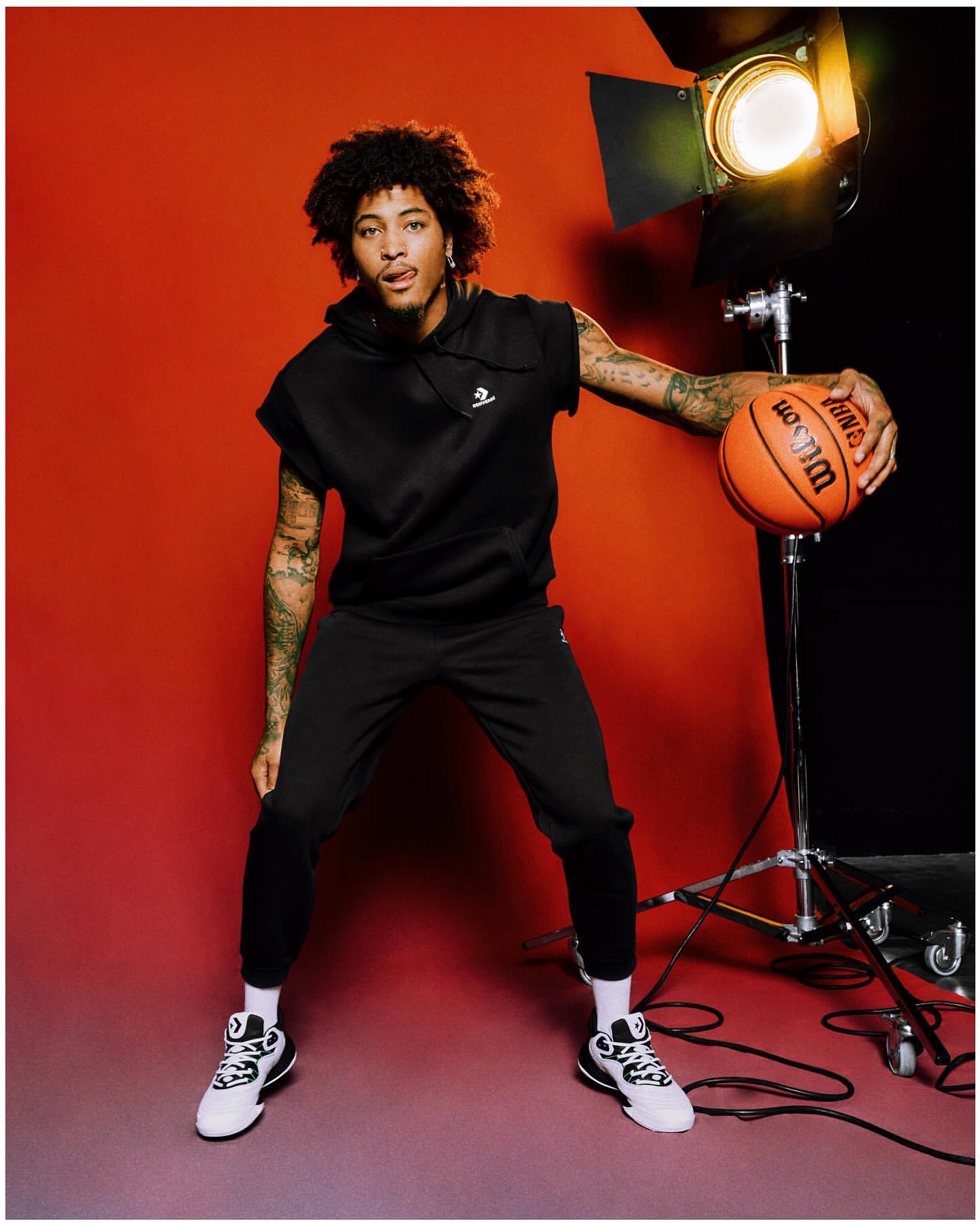 Man in black activewear poses in studio with basketball and bright lighting. He is wearing Converse shoes in a portrait shot.